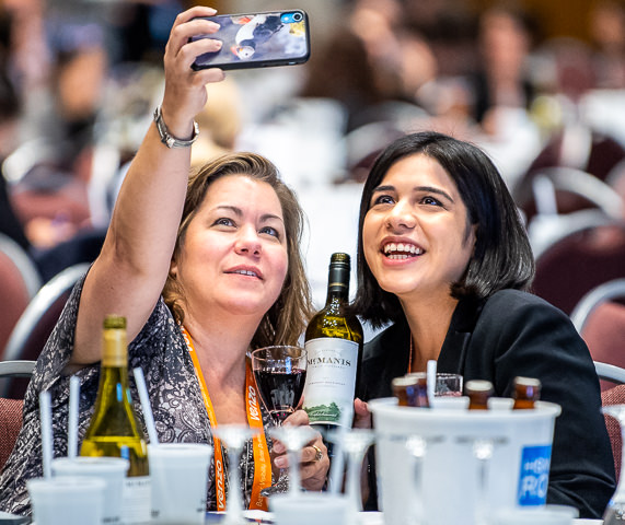 Two attendees take a selfie with a bottle of wine at a reception during an HSMAI conference at the Minneapolis Convention Center.