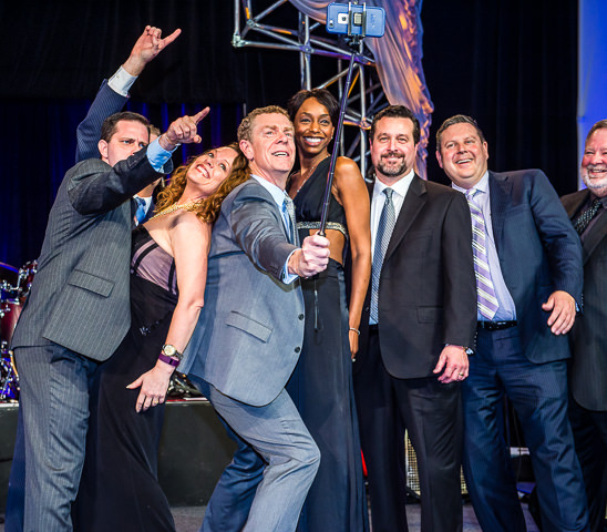 At a corporate awards ceremony, the top performing sales office poses for an on-stage selfie.